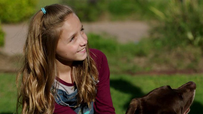 Chica rubia mirando al costado y sonriendo, con un perro marrón.