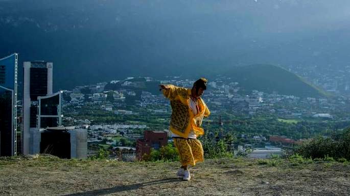 película mexicana Ja no estoy aquí. Un joven mexicano baila con ropa colorida en un escenario al aire libre con vistas a la ciudad de fondo